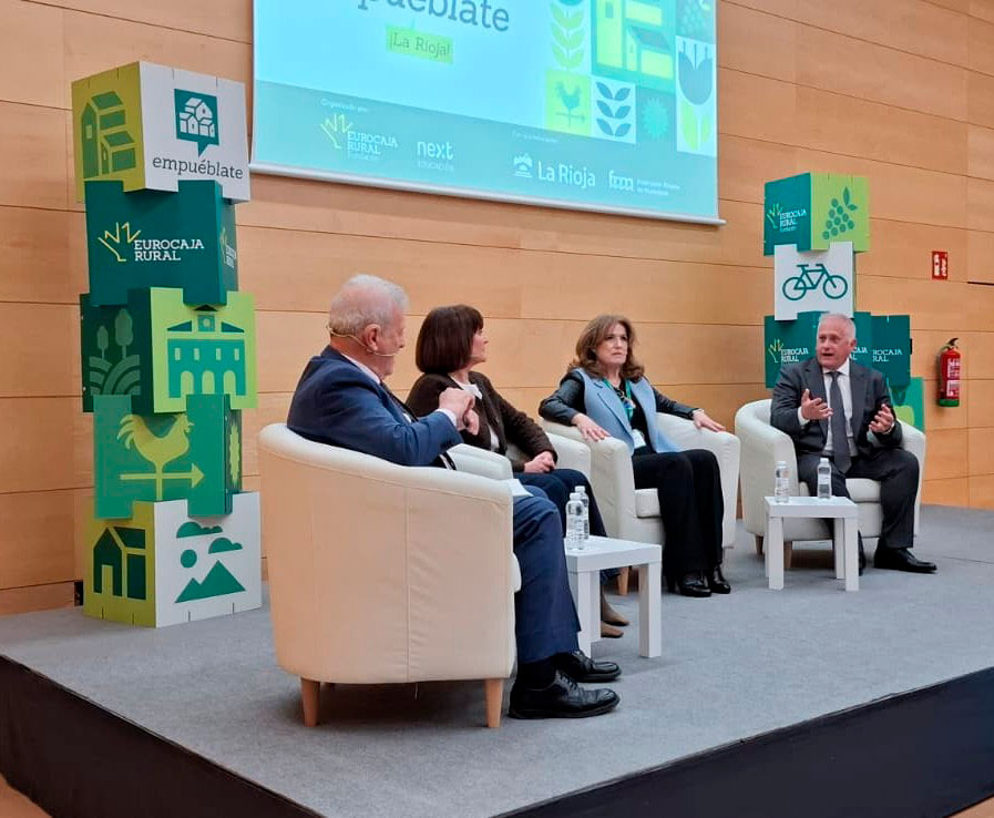 Ponencia "El papel dinamizador de los ayuntamientos" Escena de una mesa redonda en el auditorio, donde cuatro personas sentadas en sillones blancos participan en un coloquio sobre desarrollo rural. Al fondo se proyecta la imagen del evento ‘Empuéblate La Rioja’, acompañada de elementos gráficos en tonos verdes relacionados con el entorno rural. A ambos lados del escenario hay columnas decorativas con ilustraciones de naturaleza, agricultura y movilidad sostenible.”