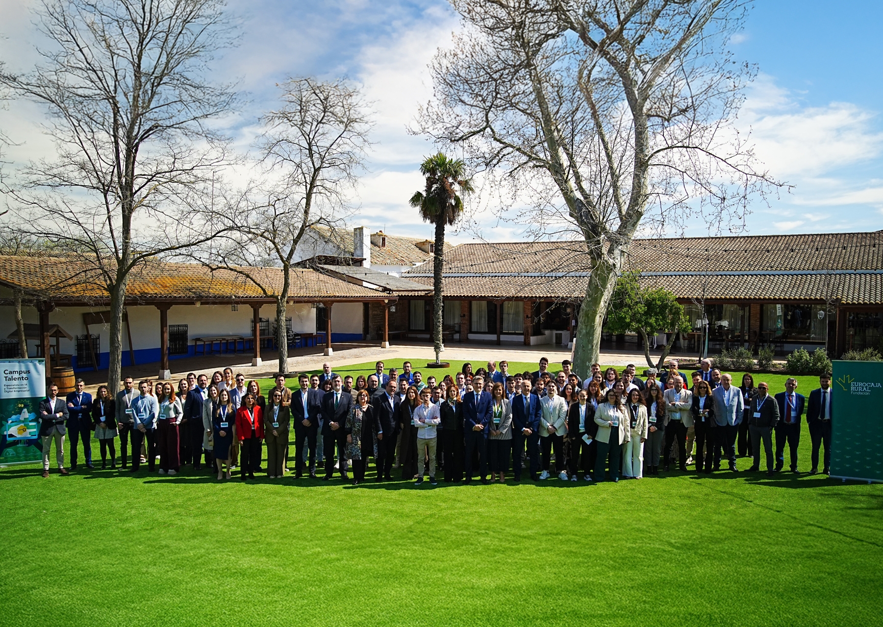 Grupo numeroso de personas posando al aire libre sobre un jardín, durante un encuentro entre empresas y talento joven del programa Campus Talento, en un espacio con edificios rurales y árboles al fondo.