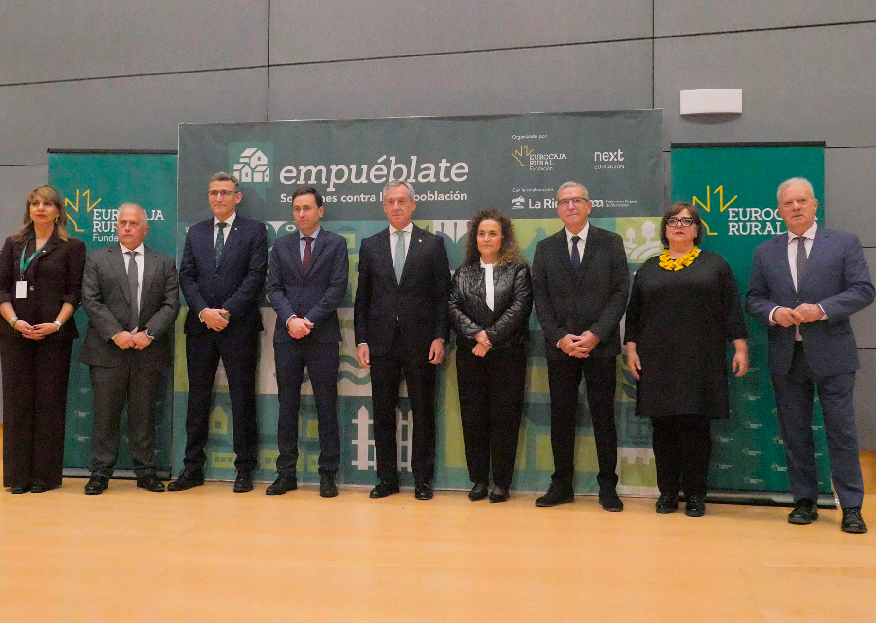 Foto de familia Empuéblate La Rioja Fotografía de un grupo de personas posando en un escenario frente a un panel del evento ‘Empuéblate’, dedicado a las soluciones contra la despoblación. Están alineadas en una fila, vestidas con indumentaria formal, con los paneles y logotipos de las entidades organizadoras y colaboradoras visibles a ambos lados y en el fondo.